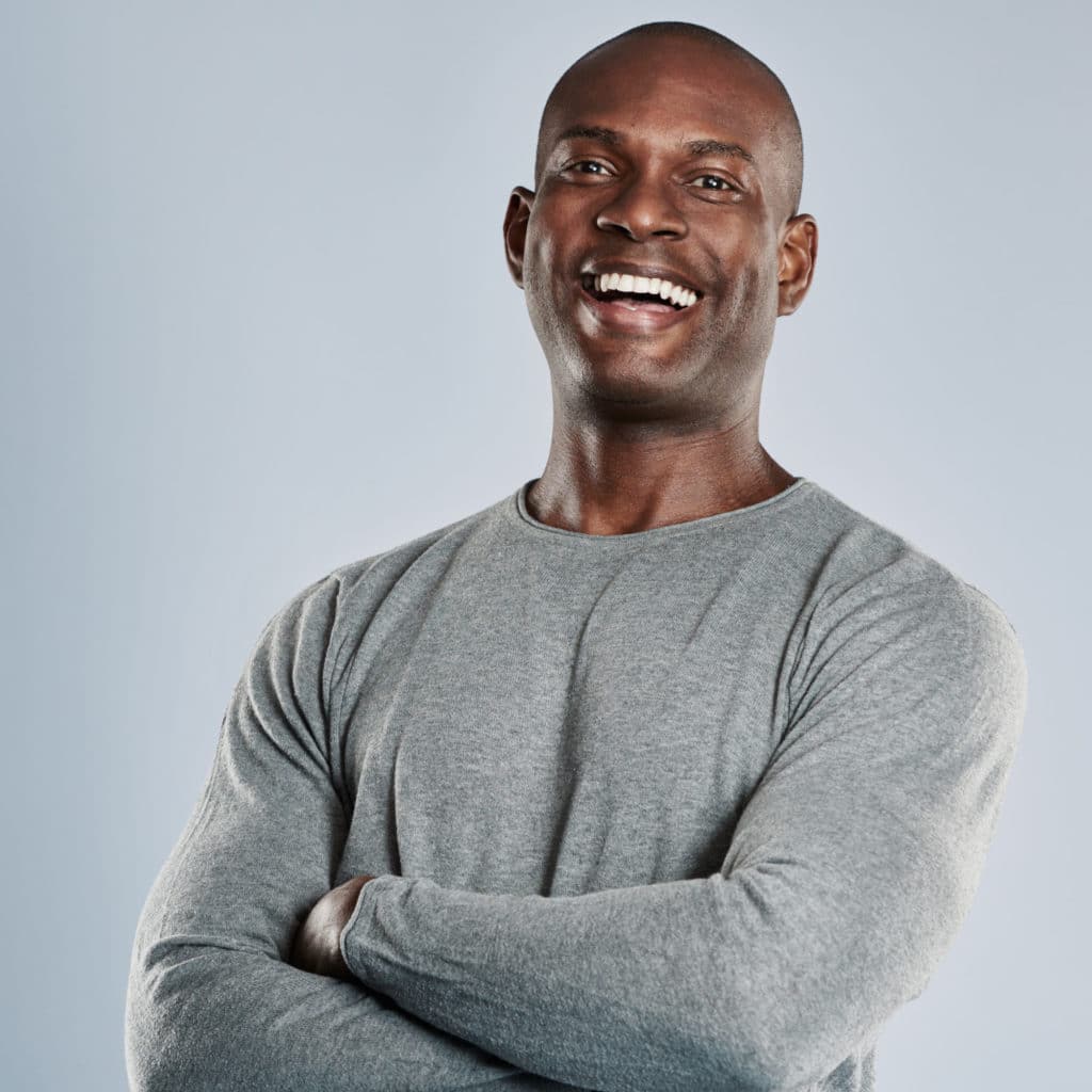 Laughing single handsome young African man with bald head in gray long sleeve shirt and folded arms over neutral background with copy space